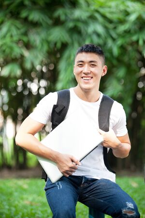 Portrait of young handsome asian student with laptop in the park, smile