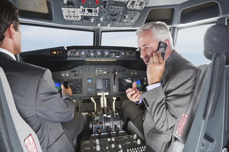 Two businessmen driving airplane in cockpit and using mobile phone at the same time