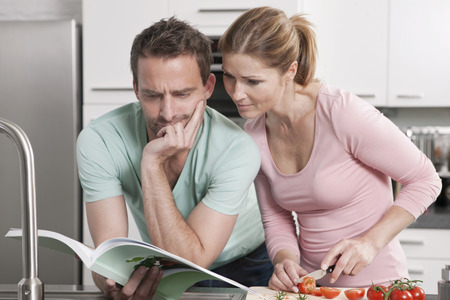 Germany, Couple reading book and preparing salad in kitchenの写真素材
