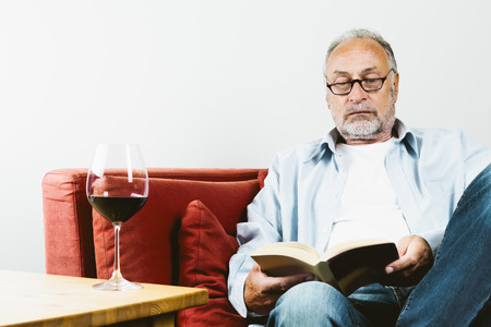 Senior man sitting on couch reading book wine glass with red wine standing on table next to himの写真素材