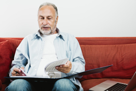 Senior man sitting on red sofa holding folder laptop next to himの写真素材