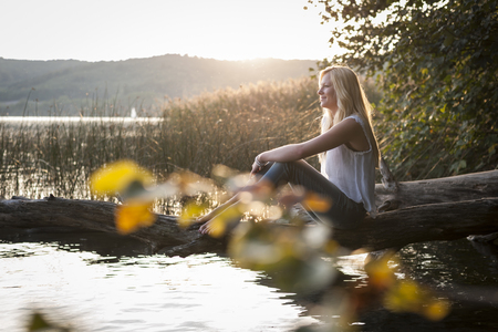 Young woman sitting on tree trunk at lakeの写真素材