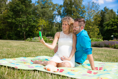 Happy couple expecting baby, sitting on blanket in parkの写真素材