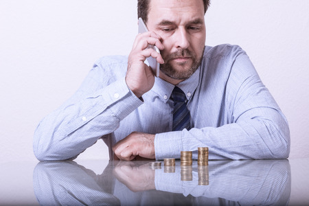 Mature man at desk with stacked euro coins using smart phoneの写真素材