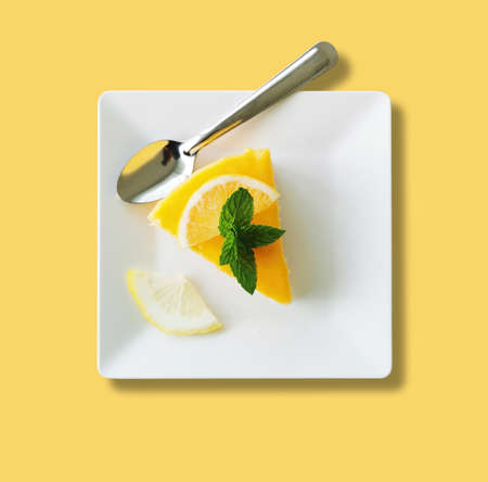 A piece of lemon cake on white square plate over yellow bachground, with the silver spoon and green mint leaves on topの写真素材