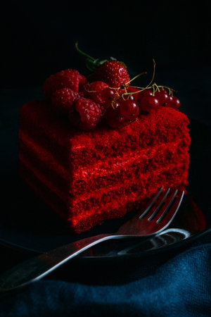 A piece of red velvet cake with raspberries and currants on a black plate with a fork on a blue...