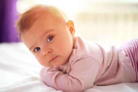 Cute three months old Baby girl infant on a bed on her belly with head up looking into camera with...