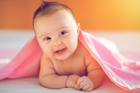 Cute three months old Baby girl infant on a bed on her belly with head up looking into camera with her big eyes. Natural bedroom light.の写真素材