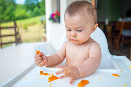 Funny adorable and dirty little baby girl of 6 months eats while sitting in the baby chair.の写真素材