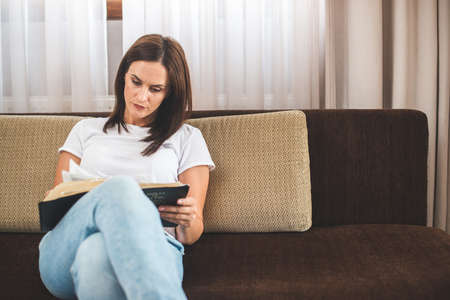 Woman Sitting On Sofa And Attentively Reading Bible. Religion Concept.の写真素材