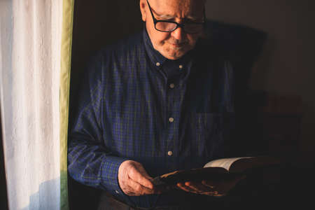 Elderly man stand next to window and reading Bibleの写真素材
