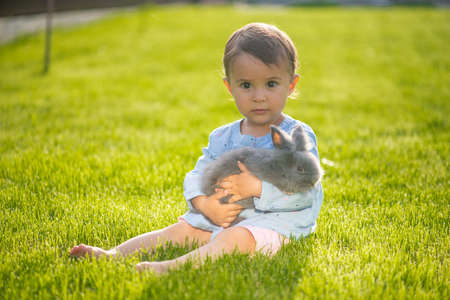 Little and cute girl hold little brown rabbit in her hands while she is sitting on grassの写真素材