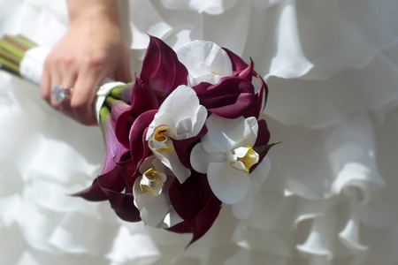 Beautiful white and dark red wedding bouquet In a female hand with a ring in white dress on backgroundの写真素材