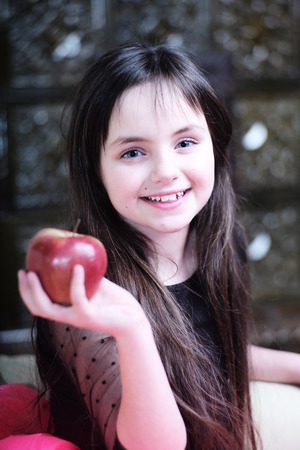 Pretty smiling little girl with red apple sitting on couch looking forward, vertical pictureの写真素材
