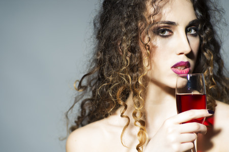 Portrait of attractive young pretty woman with curly hair and bright make up tasting red wine holding glass looking forward on light grey wall background copy space, horizontal pictureの写真素材