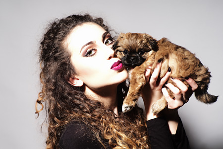 Portrait of fashionable young beautiful girl with curly hair and bright make up holding and kissing cute little brown puppy looking forward on light grey background, horizontal pictureの写真素材