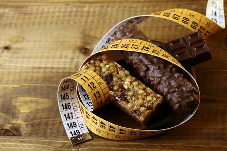 Delicious chocolate bars and peanut brittle with a measuring tape as a symbol of diet on brown wooden table top background, horizontal photoの写真素材