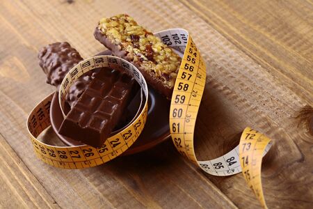 Appetizing chocolate bars and peanut brittle with a measuring tape as a symbol of diet on brown wooden table top background, horizontal photoの写真素材