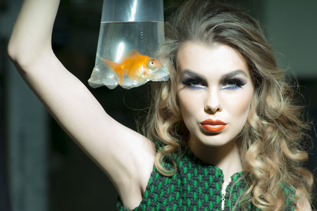 Sexy young girl with bright makeup and blonde curly hair holding cellophane package aquarium with goldfish, horizontal photoの写真素材