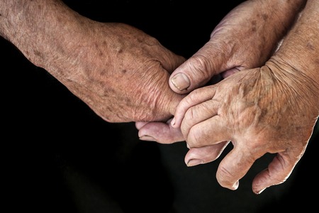 Wrinkled hands of two old elderly people holding each other closeup on black background, horizontal pictureの写真素材