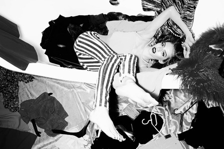 Showy bright young woman with curly hair lying in bathtub amid clothes on wall background black and white, horizontal pictureの写真素材