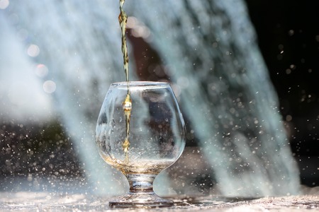 Amber alcoholic liquor poured in glass stnading on stone rim of fountain on water splashes background, horizontal pictureの写真素材