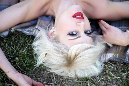Potrait of alluring sexual young blonde girl with bright makeup and red lips looking forward lying on grass on outdoor background, horizontal pictureの写真素材