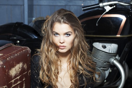 Portrait of pretty attractive young woman in black lace blouse sitting near old fashioned motorcycle with aged brown suitcase in garage on grey wooden wall background, horizontal pictureの写真素材