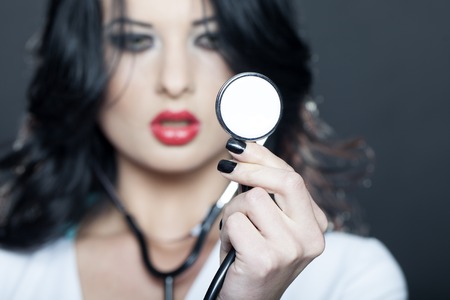Portrait of beautiful young brunette doctor with phonendoscope in white uniform standing on grey background, horizontal pictureの写真素材