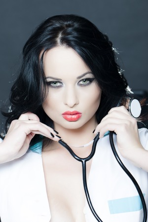 Portrait of attractive impassioned young brunette doctor with phonendoscope in white uniform standing on grey background, vertival pictureの写真素材