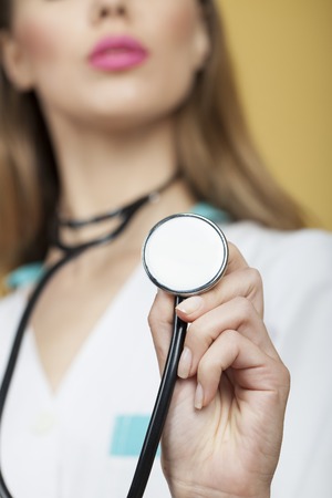 Blur closeup of young nurse in white medical uniform with focus on stethoscope on neck standing on yellow background, vertical pictureの写真素材
