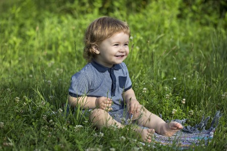 Small funny happy smiling baby boy sitting in meadow on fresh green grass with glass cup, horizontal...