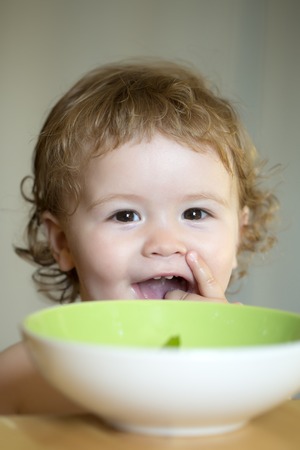 Portrait of smiling sweet little boy with blonde curly hair and round cheecks eating from green plate with hand closeup, vertical pictureの写真素材