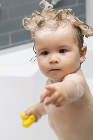 Little pretty baby boy standing in bathroom with wet foam crazy hairdo holding yellow duckling toy hold out hand, vertical pictureの写真素材