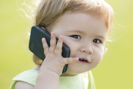 Portrait of small pretty smiling baby boy with blonde curly hair and cute face holding and speaking on black mobile phone sunny day outdoor on green grass background, horizontal pictureの写真素材
