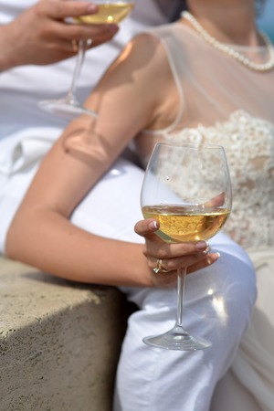 Young wedding couple of two people in white clothes holding glasses with white wine sitting and standing sunny day outdoor, vertical pictureの写真素材