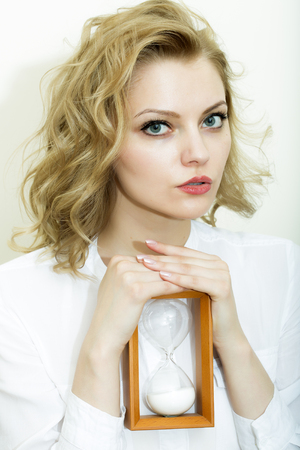 Portrait of pretty thoughtful secretary woman sitting in shirt holding wooden sand glass looking...