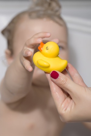 Little child sitting in bathroom with wet hair taking yellow duckling toy from hand of mother,...
