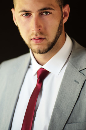 Portrait of attractive sexy unshaven businessman in grey jacket white shirt and red tie looking forward standing on black background, vertical pictureの写真素材