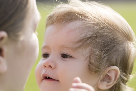 Small beautiful blond baby boy with curly hair smiling to his young mother holding him on hands...