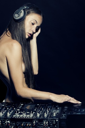 Attractive naked sexy musical young dj girl with long hair standing near mixer console with head phones with pastes on black background copyspace, vertical pictureの写真素材