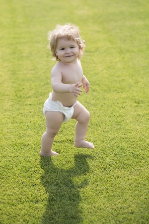 Little sweet smiling male kid with blond curly hair in white diaper making walking on lawn with fresh green grass sunny day outdoor in summer, vertical pictureの写真素材