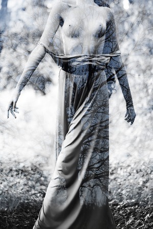 Sexy attractive young undressed woman with naked breast in long clothes standing thowing head back with bare tree branches texture on natural background black and white, vertical pictureの写真素材