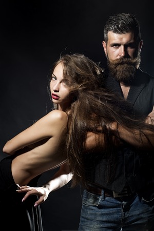 Young pair of sexual pretty girl with straight body with naked back and long hair standing near unshaven man with beard and moustache in waistcoat on black background copyspace, vertical pictureの写真素材