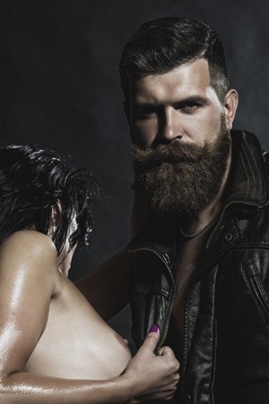 Young couple of brunette naked girl with soft skin and bare breast embracing unshaven man with beard and moustache in leather biker jacket standing on black background, vertical pictureの写真素材