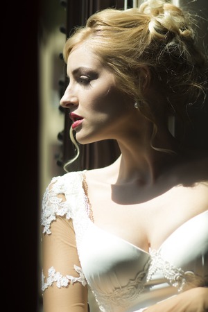 Closeup profile of beautiful tender pensive young blonde bride in white dress with elegant hairstyle standing in sunlight indoor looking away, vertical pictureの写真素材