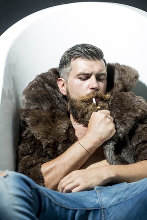 Confident unshaven man with long beard and moustache in brown fur coat with collar and blue jeans...