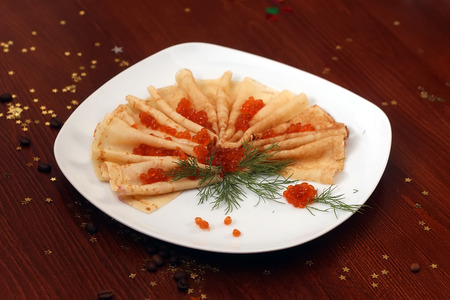 Portion of pancake with red salmon caviare above and fresh green dill on white plate on wooden table with coffee beans and decorative gold star bon-bon, horizontal pictureの写真素材