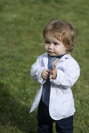 Stylish small baby boy with blond curly hair in white unbuttoned shirt grey necktie and trausers standing barefoot on fresh green grass lawn sunny day outdoor on natural background, vertical photoの写真素材