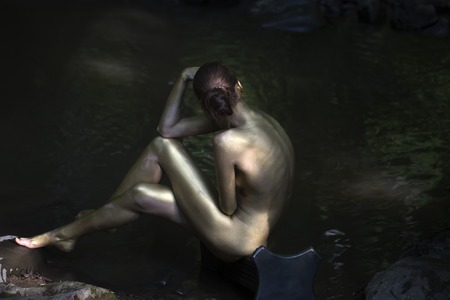 Beautiful naked woman mermaid with sexy painted gold body sitting with back on stony river coast in forest near water sunny day outdoor on natural background, horizontal pictureの写真素材